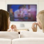 Two women in living room watching television eating chocolates a